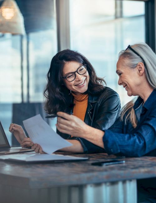 Two people sit at a table with papers and a laptop, smiling and discussing a document in a bright, modern office.