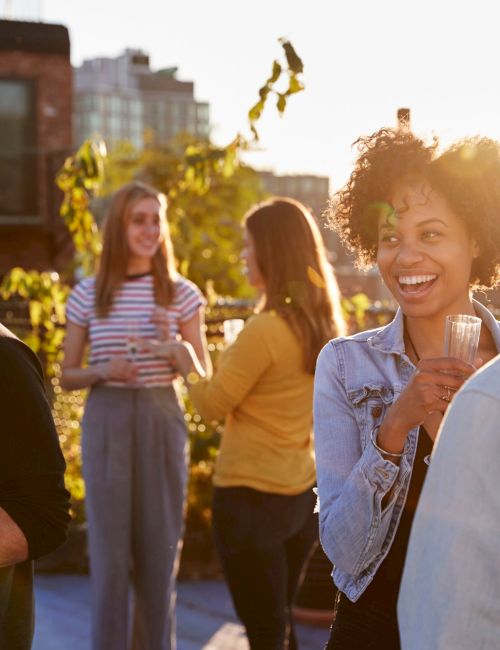 A group of friends enjoying a sunny rooftop party, chatting and holding drinks, with city buildings in the background, smiling and relaxed.
