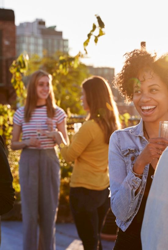 A group of young adults socializes on a sunny outdoor terrace, chatting and holding drinks, with a cityscape in the background and warm lighting.