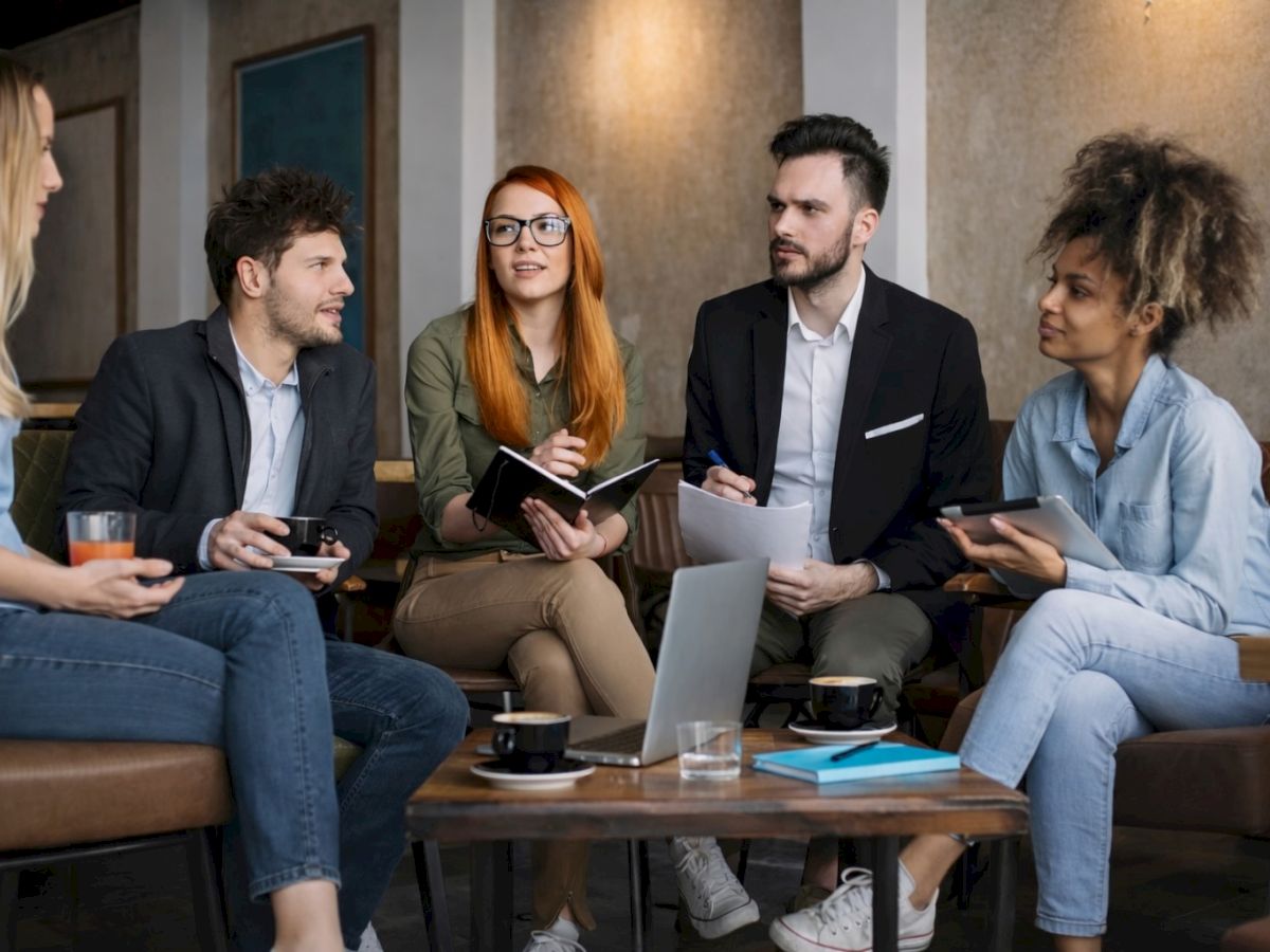 A diverse group of five friends in a casual meeting room, chatting and using laptops and tablets, with coffee on a table.