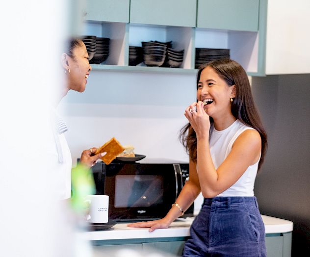 A woman in a kitchen grabbing a snack from the fridge, wearing a tank top and jeans, with shelves and food visible inside.