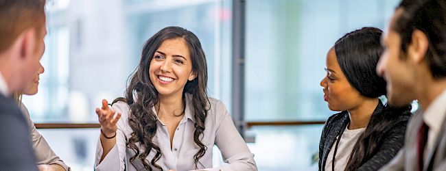 Three people in a meeting around a table with papers, a laptop, and a cup; one person smiles as others discuss, in a bright office.