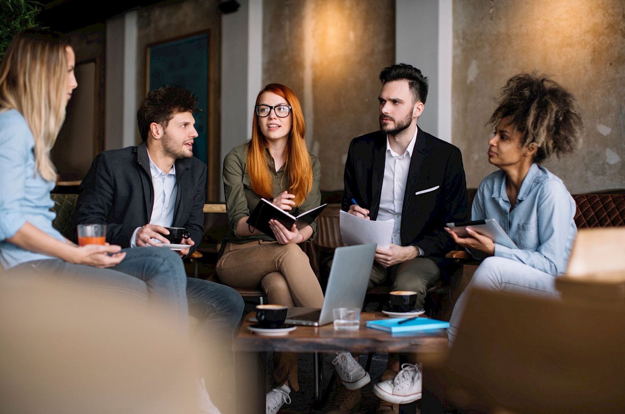 A group of five young professionals sit in a modern lounge, sharing a discussion with laptops, notebooks, and phones in hand, collaborating intently.