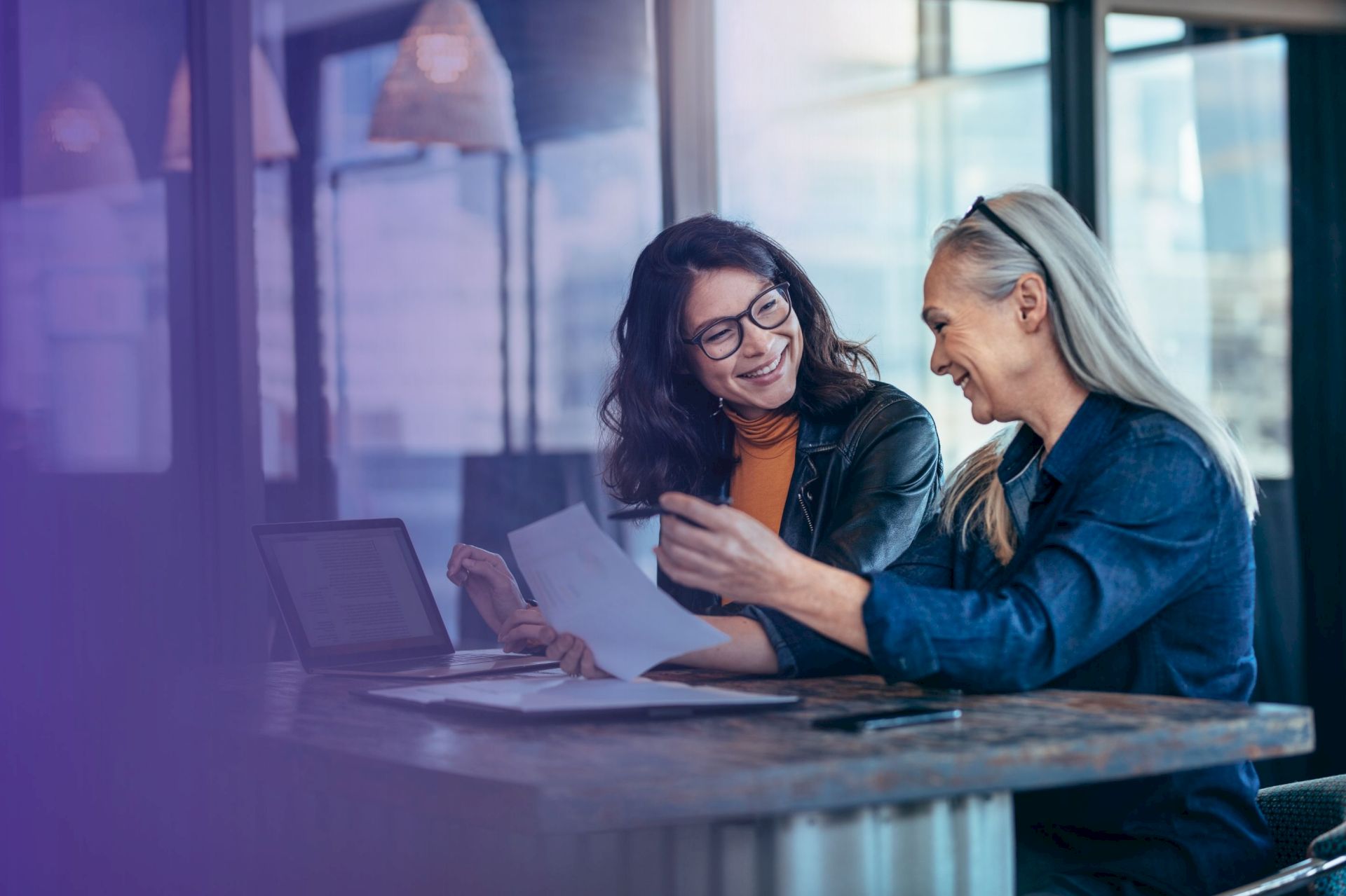 Two colleagues smile over papers at a desk with a laptop, sharing ideas in a bright office meeting.