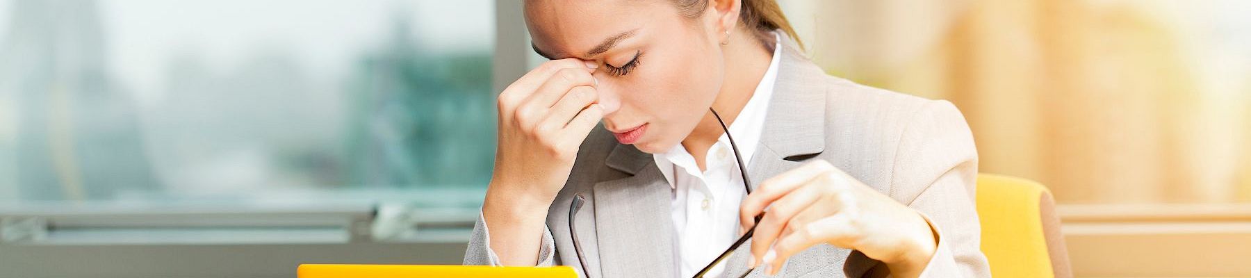 A tired businesswoman in a beige blazer sits at a desk, pinching the bridge of her nose, holding glasses, with a yellow tablet in view.