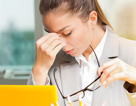 A tired businesswoman in a beige blazer sits at a desk, pinching the bridge of her nose, holding glasses, with a yellow tablet in view.