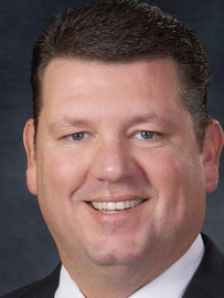 A smiling man in a dark suit and white shirt, head and shoulders portrait, neutral studio background. Top it at 140 characters, always ending the sentence.
