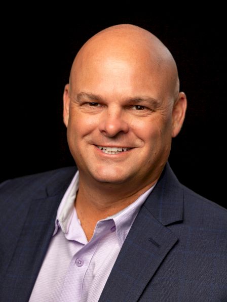 A smiling bald man in a navy blazer and light purple shirt poses for a portrait, set against a dark background.