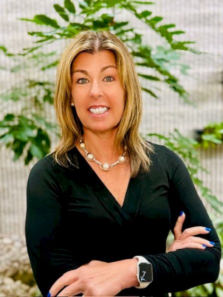 A confident woman with blond hair in a black V-neck top, wearing a pearl necklace and a smartwatch, standing with arms crossed outdoors.