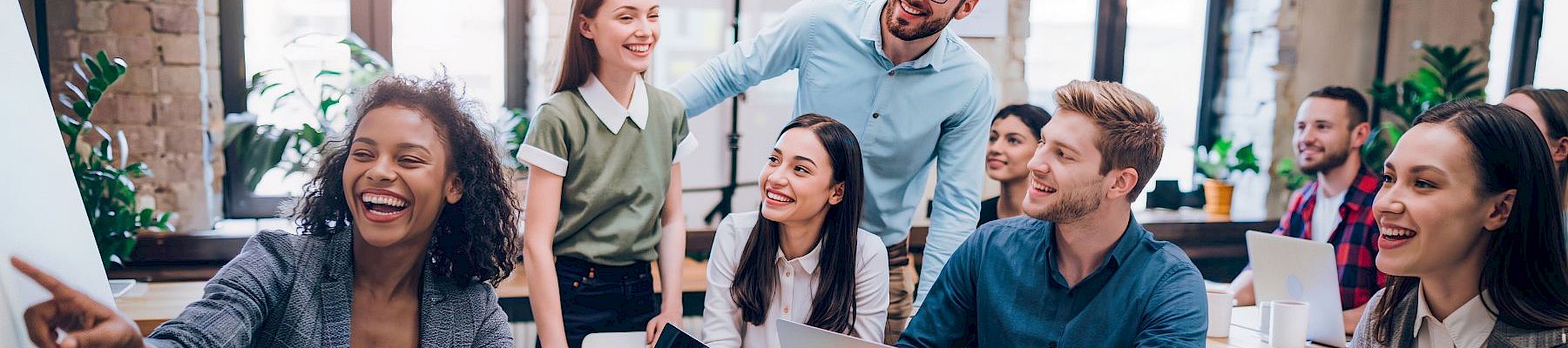 A diverse group of coworkers in a bright office are collaborating around a table with laptops, laughing and sharing ideas.
