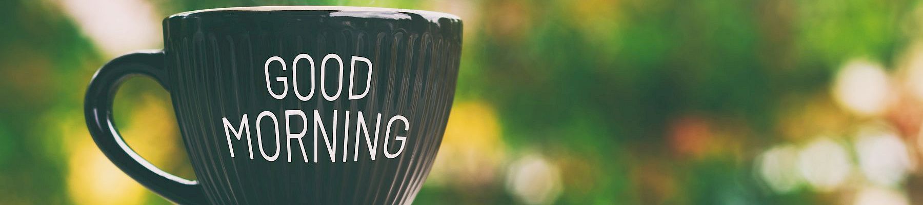 A black mug with &ldquo;GOOD MORNING&rdquo; in white letters sits on a surface, blurred greenery in the background, radiating cheerful morning vibes.