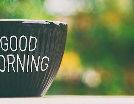 A black mug with &ldquo;GOOD MORNING&rdquo; in white letters sits on a surface, blurred greenery in the background, radiating cheerful morning vibes.
