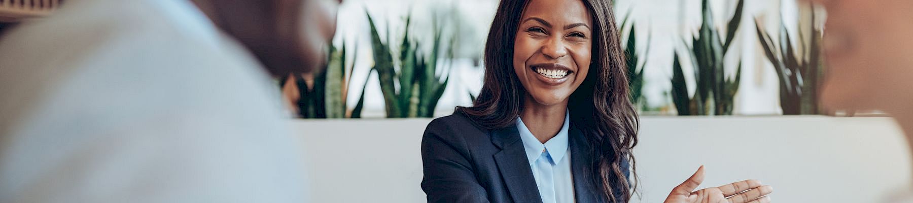 A friendly business meeting in progress, a smiling woman in a blazer greets colleagues with an open, welcoming gesture.
