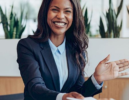 A friendly business meeting in progress, a smiling woman in a blazer greets colleagues with an open, welcoming gesture.
